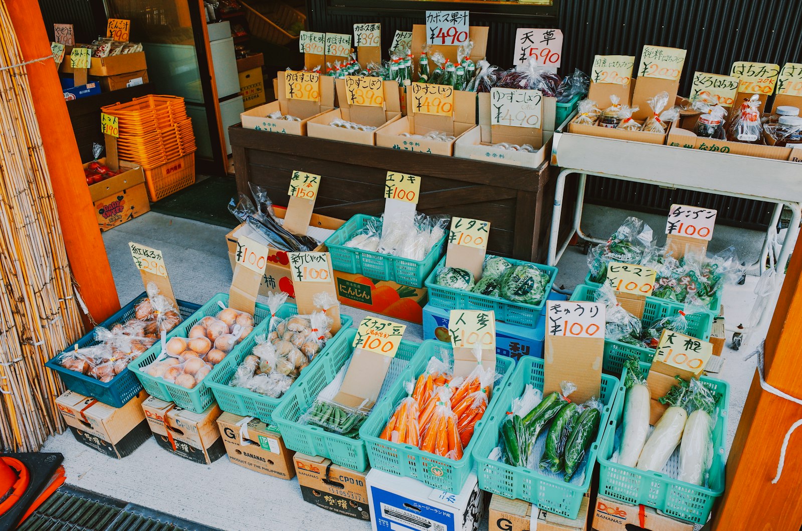 The front of a farm stand, with vegetables lined up in plastic containers and cardboard boxes and hand-written price tags standing among them